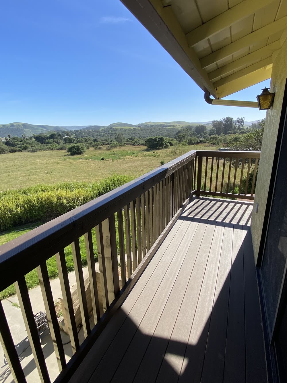 A second-story deck with a brown wood railing looking out over open grassland and green hills.