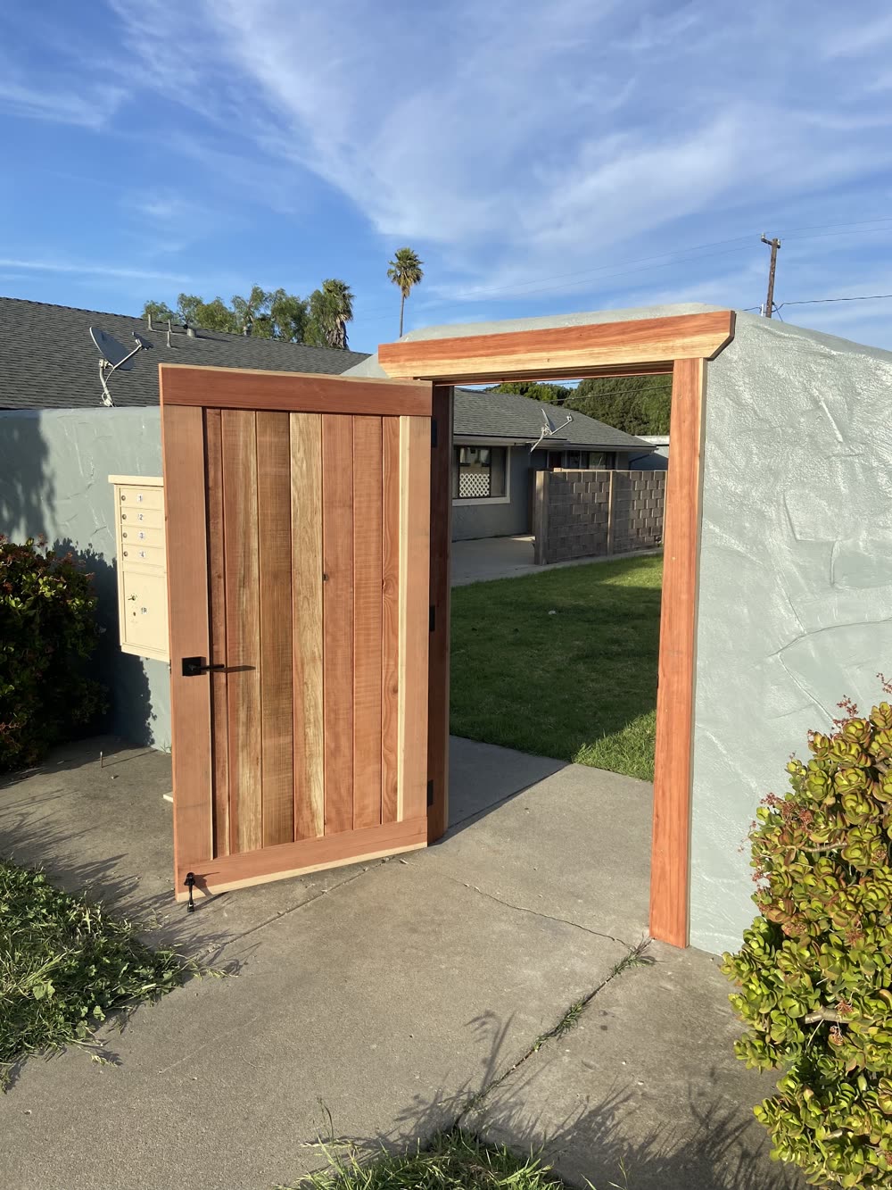 A new redwood gate standing open in a redwood frame set into a gray stucco wall, with a lawn and an apartment building beyond.