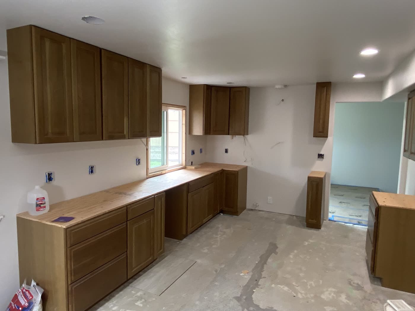 A kitchen with oak cabinets set on the walls and floor, plywood counter tops in place and bare concrete underfoot.