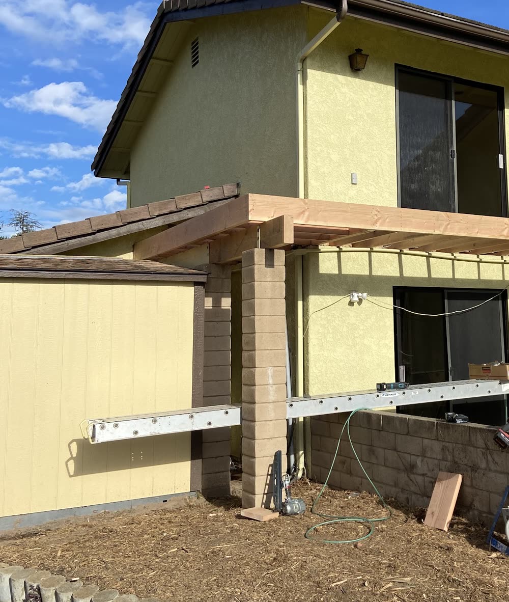 A timber patio cover going up on block columns against a yellow stucco house, with an aluminum plank and tools on the ground.